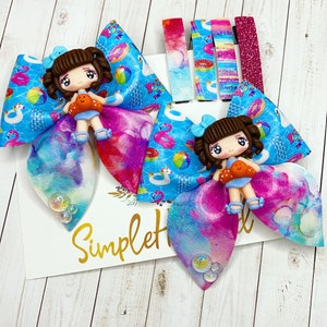 May include: Two colorful hair bows with a summer theme. Each bow features a blue and white patterned fabric with images of inflatable pool toys, a beach ball, and a unicorn. A small doll with brown hair and a blue dress is attached to each bow. The bows are tied with a pink and blue tie-dye ribbon.