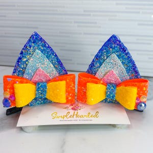 May include: A pair of handmade hair accessories featuring glittery cat ear shapes in blue, light blue, and pink, with orange and yellow bows. The accessories are embellished with iridescent gems and are attached to black clips.