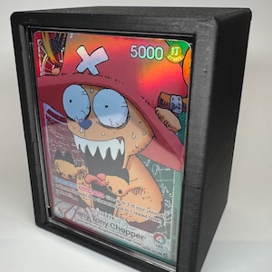 May include: A black card display case containing a holographic trading card. The card shows a cartoon character wearing a red hat and the text "5000 STRIKE". The case is made of black plastic.