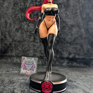 May include: A detailed figurine of a woman with red hair, wearing a black outfit, including a top, gloves, and thigh-high stockings. The figure stands on a black base with a red emblem. A sticker with a colorful design is visible.
