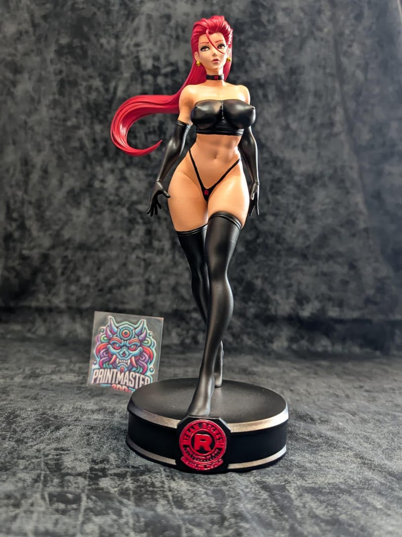 May include: A detailed figurine of a woman with red hair, wearing a black outfit, including a top, gloves, and thigh-high stockings. The figure stands on a black base with a red emblem. A sticker with a colorful design is visible.
