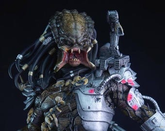 May include: A detailed Predator alien figurine, showcasing a textured head with open jaws and sharp teeth. The figure is clad in metallic armour with red accents and intricate details, including dreadlocks and shoulder-mounted weaponry. The colour scheme is a mix of greens, browns, and silvers.