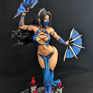 Puede incluir: Figura detallada de un personaje femenino con un traje azul, sosteniendo abanicos azules. La figura tiene cabello negro, una máscara facial y detalles intrincados en el atuendo. La base presenta calaveras y detalles rojos.