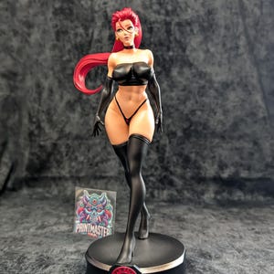 May include: A detailed figurine of a woman with red hair, wearing a black outfit, including a strapless top, gloves, and thigh-high stockings. The figure stands on a round black base with a red logo. A sticker with colorful artwork is visible.