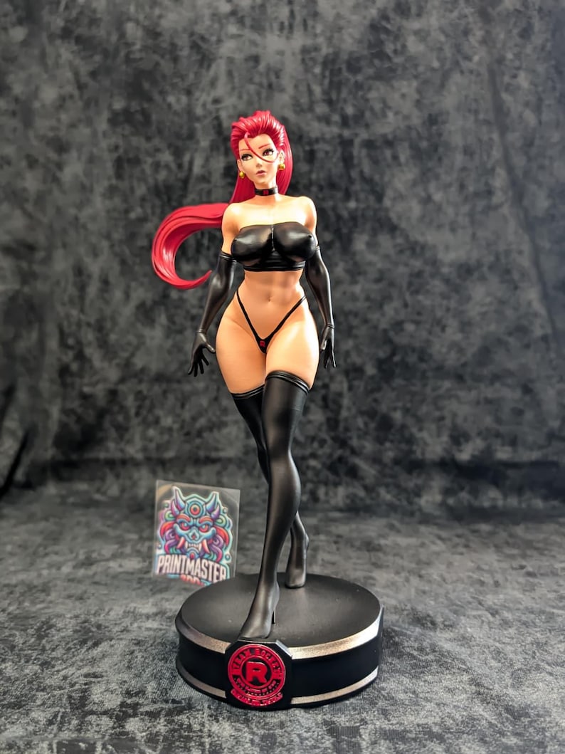 May include: A detailed figurine of a woman with red hair, wearing a black outfit, including a strapless top, gloves, and thigh-high stockings. The figure stands on a round black base with a red logo. A sticker with colorful artwork is visible.