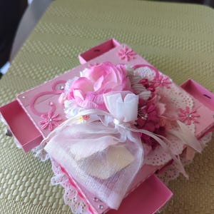 May include: A pink box with a white lace trim and a pink flower arrangement on top. The box has multiple compartments and is decorated with rhinestones.