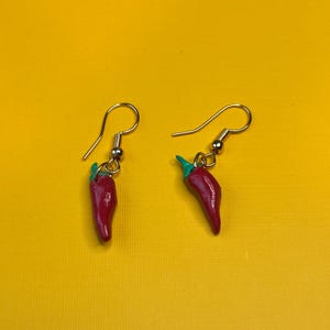 Könnte beinhalten: Ein Paar handgefertigte Ohrringe mit roten Chili-Pfeffer-Anhängern mit grünen Stielen. Die Ohrringe hängen an goldfarbenen Fischhaken-Ohrdrähten. Der Hintergrund ist einfarbig gelb.