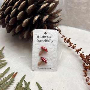 May include: A pair of small mushroom-shaped stud earrings with red caps and white spots on a white rectangular display card. The card features the text "francifully" and "francifully.co.uk" along with small icons of a pinecone, moon, butterfly, and mushroom.