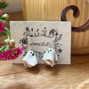 Op de afbeelding: Handgemaakte spookoorbellen. Elke oorbel is een klein, wit spookje met zwarte stipogen en een gebogen basis, hangend aan zilverkleurige haken. De oorbellen worden gepresenteerd op een kaart met het woord "francifully" erop. De achtergrond bevat roze bloemen.