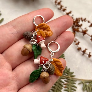 May include: A pair of silver hoop earrings featuring dangling charms shaped like autumn elements, including a red and white mushroom, a brown acorn, a textured pinecone, a green leaf, and a golden-yellow maple leaf, all crafted with detail.