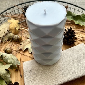 May include: A light blue geometric candle with a wick, resting on a beige linen cloth. The candle is surrounded by fall foliage, including leaves, acorns, and pinecones, on a wooden surface.
