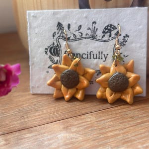 Puede incluir: Pendientes de girasol hechos a mano con ganchos dorados. Los pendientes presentan pétalos amarillos y un centro marrón. La palabra "ncifully" es visible en la tarjeta blanca detrás de los pendientes. Un accesorio encantador.