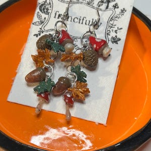 Puede incluir: Pendientes colgantes con bellotas, setas y hojas en miniatura. Los pendientes tienen ganchos plateados y se exhiben en una tarjeta blanca dentro de un plato naranja. Las setas tienen gorros rojos con lunares blancos.