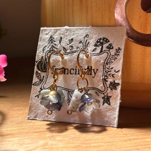 Può includere: Un paio di orecchini a cerchio color oro con delicati ciondoli floreali blu e bianchi traslucidi. Gli orecchini sono appesi davanti a una carta bianca con la parola "fancily" e illustrazioni decorative. Lo sfondo è una superficie di legno.