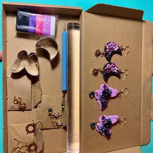 May include: A craft kit with polymer clay, tools, and earring components. The kit includes black, purple, and white clay, a roller, and various earring designs. Finished earrings feature floral and geometric designs in black, purple, and gold.