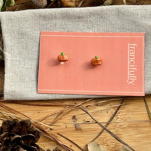 May include: Two pumpkin-shaped stud earrings with green stalks, presented on a pink card with the word "francifully". The card sits on a grey linen pouch, with a wooden background and autumn leaves.