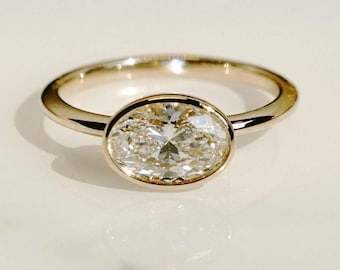 Può includere: Un anello d'oro con un grande diamante taglio ovale. Il diamante è incastonato in una montatura a castone e la fascia è in oro lucido semplice. L'anello è esposto su una superficie bianca, che riflette la luce.