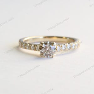 Puede incluir: Un anillo de compromiso de oro con un gran diamante redondo y transparente en el centro. La banda también está adornada con diamantes más pequeños, creando un efecto brillante. El anillo se encuentra sobre un fondo suave y neutro.