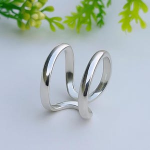 Könnte beinhalten: Ein silberfarbener Ring mit einem einzigartigen Doppelband-Design. Der Ring hat zwei gebogene Bänder, die sich an der Basis vereinen und eine moderne und minimalistische Ästhetik schaffen. Der Ring ist vor einem weißen Hintergrund mit grünem Laub platziert.