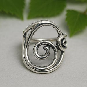 May include: A silver ring with a swirling spiral design. The ring features a large, open spiral in the centre, with a smaller spiral detail on the side. The ring is set against a light grey background with green leaves.