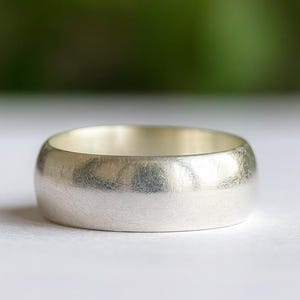 Pode incluir: Um anel de banda de prata com um acabamento escovado. O anel tem uma forma arredondada e um interior liso e polido. O anel está sobre uma superfície branca com um fundo verde desfocado.