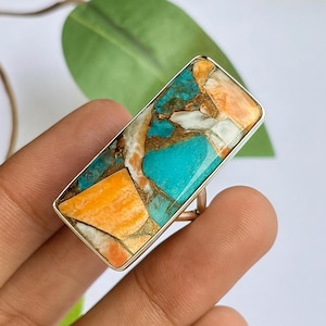 May include: A rectangular ring with a silver band and a large, colorful stone. The stone features a mosaic of turquoise, orange, white, and gold-colored sections. The ring is set against a neutral background, highlighting the vibrant gemstone.