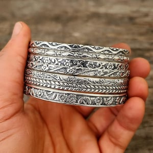 May include: A collection of silver bangles, each adorned with detailed floral and leaf motifs. The bangles are stacked, highlighting the variety of designs. The metal has a slightly aged look, adding to the vintage appeal. The bangles are held in a hand.