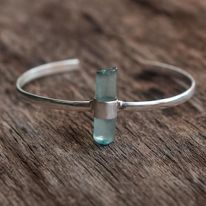 May include: A silver-toned cuff bracelet featuring a light blue, rectangular crystal centerpiece. The crystal is vertically oriented and secured by a silver band. The bracelet has a simple, open design.