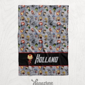 May include: A gray and black fleece blanket with a repeating pattern of small cartoon superheroes. The blanket has a black band with a red border and the name "HOLLAND" in white text. A small cartoon Iron Man is to the left of the name.