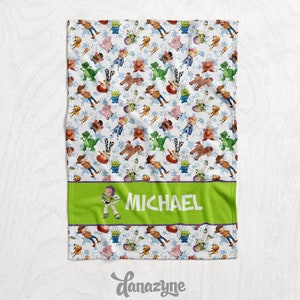 Może przedstawiać: Biały koc z polaru z zieloną obw&oacute;dką i zielonym panelem przedstawiającym kresk&oacute;wkową postać Buzz Lightyeara i imię "MICHAEL". Koc jest pokryty powtarzającym się wzorem postaci z Toy Story, w tym Buzz Lightyear, Woody, Jessie, Rex i Pan Potato Head.