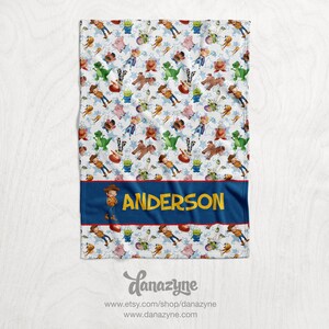 Może przedstawiać: Biały koc z polaru z powtarzającym się wzorem postaci z Toy Story, w tym Woody, Buzz Astral i Rex. Koc ma niebieską i czerwoną obw&oacute;dkę z imieniem "ANDERSON" w ż&oacute;łtych literach.