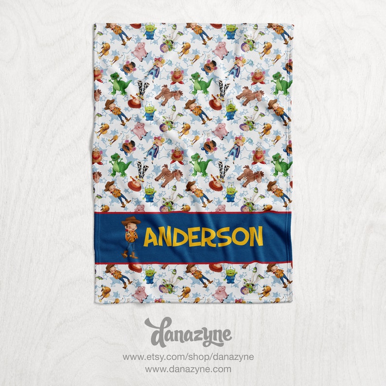 Może przedstawiać: Biały koc z polaru z powtarzającym się wzorem postaci z Toy Story, w tym Woody, Buzz Astral i Rex. Koc ma niebieską i czerwoną obw&oacute;dkę z imieniem "ANDERSON" w ż&oacute;łtych literach.