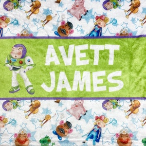 Może przedstawiać: Personalizowany koc w kolorze zielonym i fioletowym na białym tle. Koc przedstawia powtarzający się wz&oacute;r postaci z Toy Story, w tym Buzz Astral, Woody i Jessie. Imię "AVETT JAMES" jest nadrukowane białymi literami na zielono-fioletowym pasku.