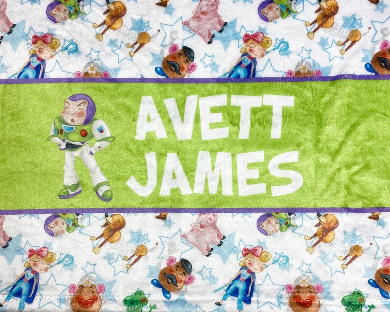 Może przedstawiać: Personalizowany koc w kolorze zielonym i fioletowym na białym tle. Koc przedstawia powtarzający się wz&oacute;r postaci z Toy Story, w tym Buzz Astral, Woody i Jessie. Imię "AVETT JAMES" jest nadrukowane białymi literami na zielono-fioletowym pasku.