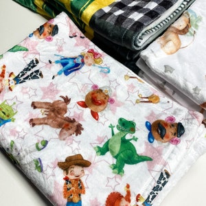 Może przedstawiać: Biały koc z polaru z wzorem r&oacute;żowych gwiazdek i kolorowymi postaciami z kresk&oacute;wek z serii film&oacute;w Toy Story. Postacie to Woody, Buzz Astral, Jessie, Rex i Pan Potato.