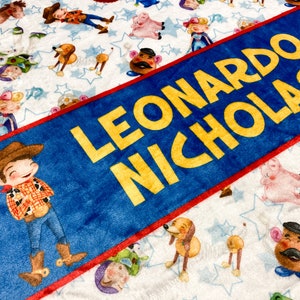 Może przedstawiać: Personalizowany koc w kolorze niebieskim i czerwonym z imionami Leonardo Nicholas w ż&oacute;łtych literach. Koc ma białe tło z powtarzającym się wzorem postaci z Toy Story.