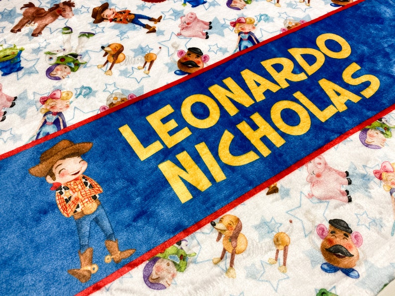 Może przedstawiać: Personalizowany koc w kolorze niebieskim i czerwonym z imionami Leonardo Nicholas w ż&oacute;łtych literach. Koc ma białe tło z powtarzającym się wzorem postaci z Toy Story.