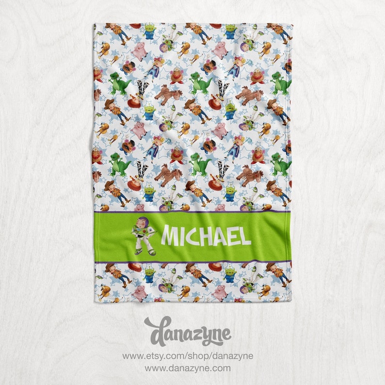 Może przedstawiać: Biały koc z polaru z zieloną obw&oacute;dką i zielonym panelem przedstawiającym kresk&oacute;wkową postać Buzz Lightyeara i imię "MICHAEL". Koc jest pokryty powtarzającym się wzorem postaci z Toy Story, w tym Buzz Lightyear, Woody, Jessie, Rex i Pan Potato Head.