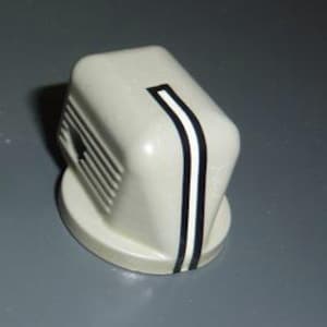 Puede incluir: Un mando de estilo retro de color crema con una franja negra y blanca en la parte delantera. El mando tiene una forma rectangular redondeada con una base circular y líneas horizontales en un lado.