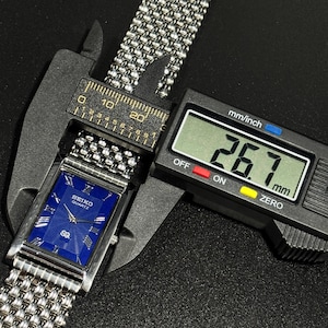 Może przedstawiać: Srebrny zegarek Seiko Quartz z niebieską tarczą i bransoletą z siatki jest mierzony suwmiarką cyfrową. Tarcza zegarka jest prostokątna ze srebrnymi wskaz&oacute;wkami i znacznikami godzin. Suwmiarka wskazuje 26,7 mm.