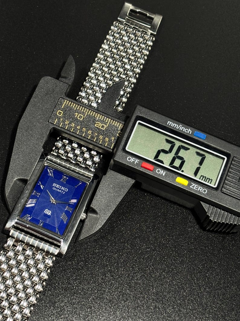 Może przedstawiać: Srebrny zegarek Seiko Quartz z niebieską tarczą i bransoletą z siatki jest mierzony suwmiarką cyfrową. Tarcza zegarka jest prostokątna ze srebrnymi wskaz&oacute;wkami i znacznikami godzin. Suwmiarka wskazuje 26,7 mm.