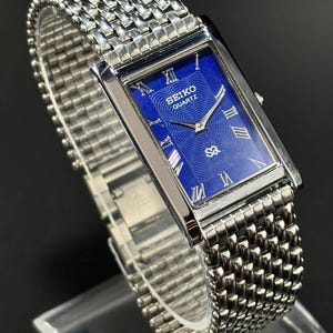 Może przedstawiać: Prostokątny zegarek Seiko Quartz z niebieską tarczą i srebrnymi rzymskimi cyframi. Zegarek ma srebrną bransoletę z siatki i srebrną kopertę. Zegarek jest prezentowany na przezroczystym stojaku.