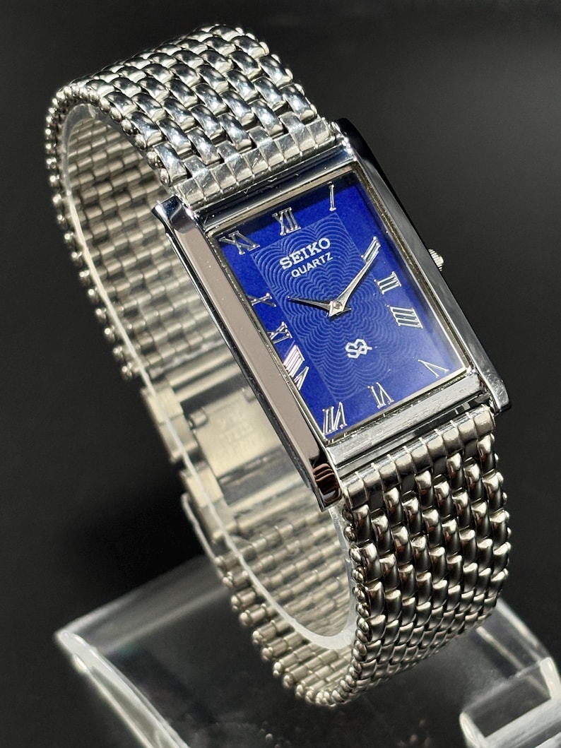 Może przedstawiać: Prostokątny zegarek Seiko Quartz z niebieską tarczą i srebrnymi rzymskimi cyframi. Zegarek ma srebrną bransoletę z siatki i srebrną kopertę. Zegarek jest prezentowany na przezroczystym stojaku.