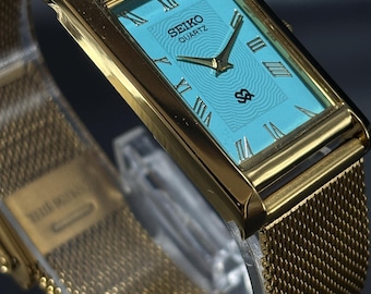 May include: A gold-toned Seiko Quartz watch with a rectangular face and a mesh band. The watch face is a light blue with gold hands and Roman numerals. The watch has the Seiko Quartz logo.