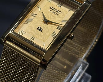 May include: A gold-toned Seiko Quartz watch with a rectangular face and mesh band. The watch face features Roman numerals and the Seiko logo. The watch is displayed on a clear stand against a dark background.