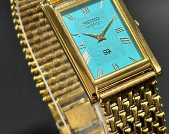 May include: A gold-toned Seiko Quartz watch with a rectangular turquoise face and gold Roman numerals. The watch features a gold mesh band and the Seiko Quartz logo. The watch is a classic design.