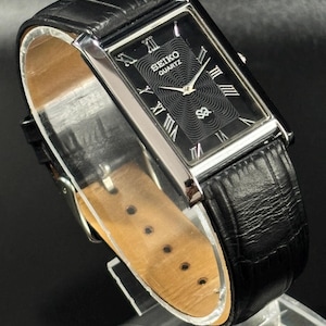 Puede incluir: Un reloj Seiko Quartz rectangular con esfera negra y caja plateada. El reloj presenta n&uacute;meros romanos y una correa de cuero negro. La esfera tiene un dise&ntilde;o texturizado y el bisel es plateado.