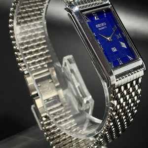 Może przedstawiać: Srebrny zegarek Seiko Quartz z prostokątną niebieską tarczą. Zegarek posiada rzymskie cyfry jako znaczniki godzin i bransoletę w stylu siatki. Zegarek jest prezentowany na przezroczystym stojaku.