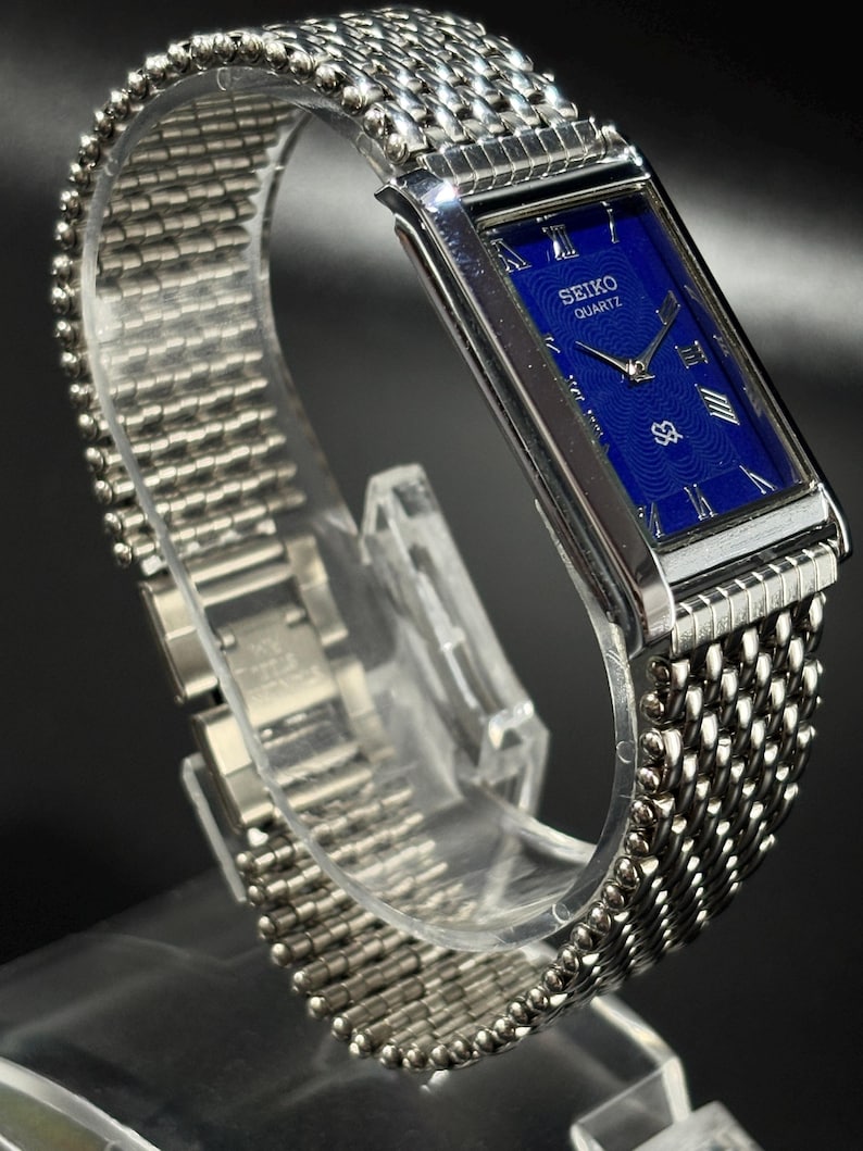 Może przedstawiać: Srebrny zegarek Seiko Quartz z prostokątną niebieską tarczą. Zegarek posiada rzymskie cyfry jako znaczniki godzin i bransoletę w stylu siatki. Zegarek jest prezentowany na przezroczystym stojaku.
