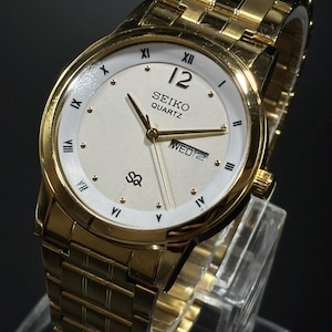 May include: A gold-toned Seiko Quartz watch with a white face, gold hands, and a gold link band. The watch face displays the day and date, with Roman numerals and gold markers. The watch is displayed on a clear stand.